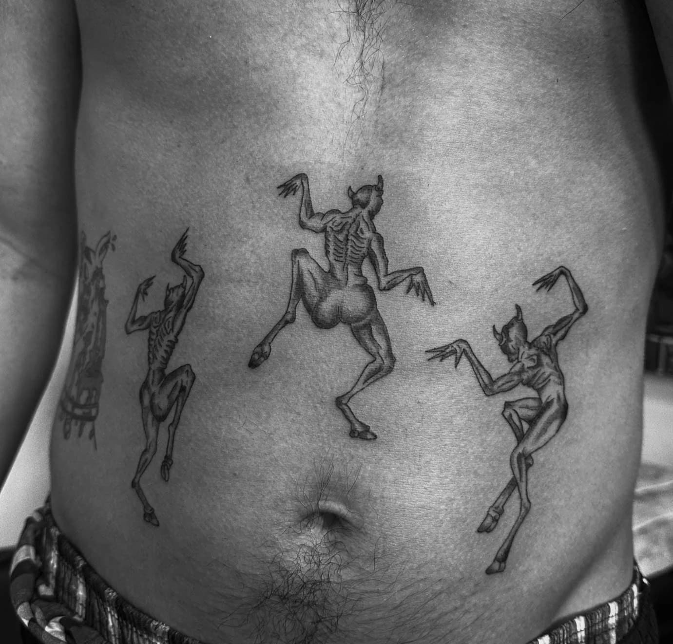 Drei surreal tanzende Figuren als feines Schwarz-Grau-Tattoo auf dem Bauch
