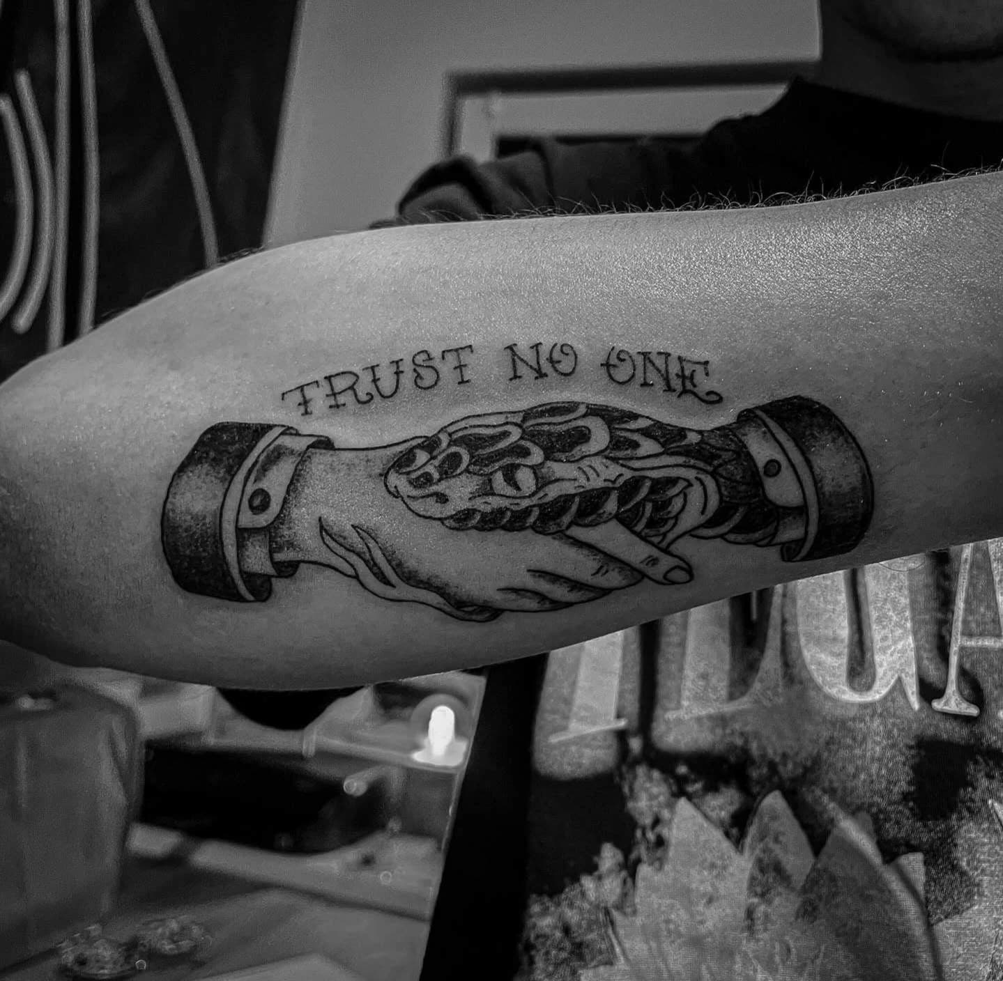 Schwarz-graues Unterarmtattoo zweier Hände unter dem Schriftzug Trust No One